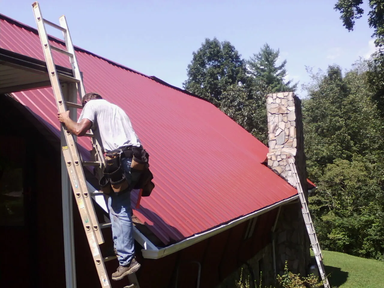 Roofer on a ladder beside a red metal roof and stone chimney — Soffit & Fascia Repair by Roofing Brothers in East Hartland